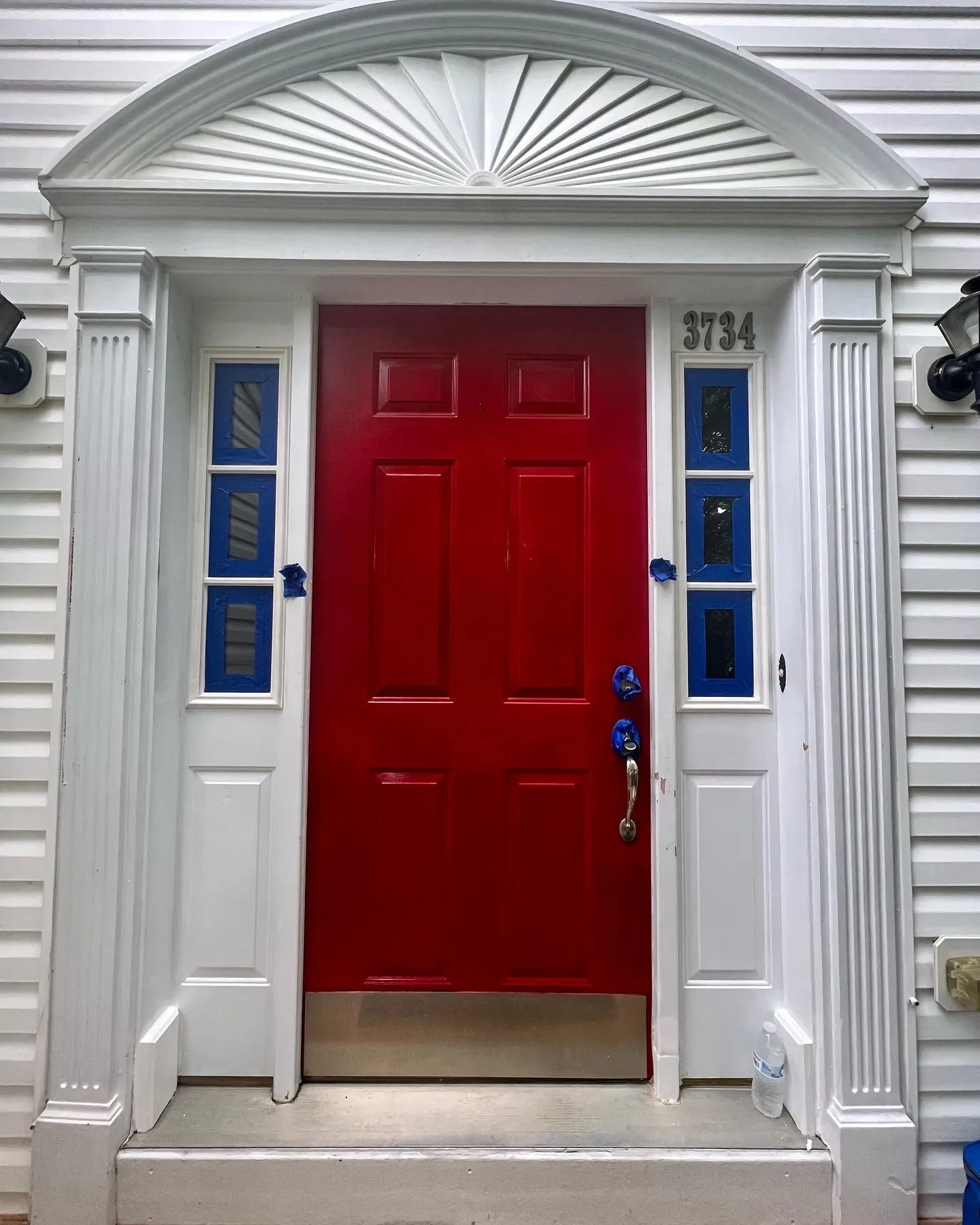 Same front door before refinish — bright red 6-panel under sunburst pediment, blue painters tape masking sidelight glass
