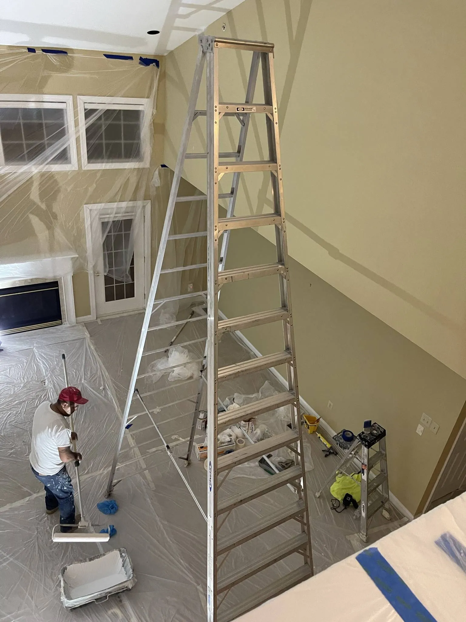 Cathedral great room post-paint, 12-ft Werner ladder centered in the space, plastic sheeting protecting the floor and the fireplace, JL crew member in red cap with paint-splattered jeans