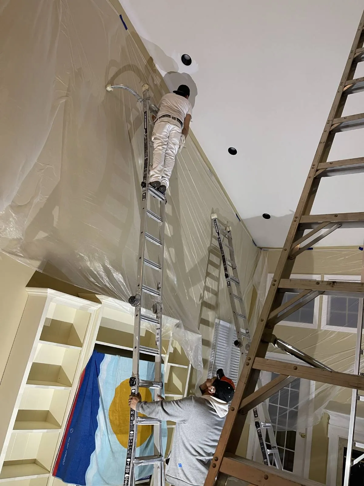 JL painter in white coveralls finishing a vaulted residential ceiling around recessed lights, plastic sheeting protecting built-in shelving below