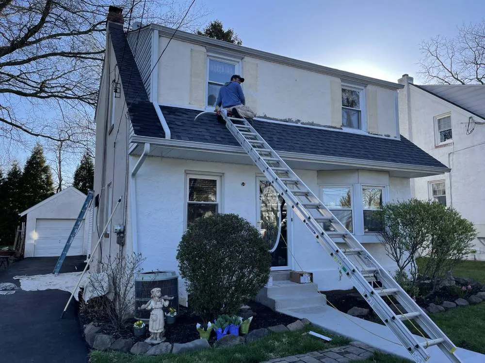 Jose Lopez on the roof of a 2-story Pennsylvania colonial mid-paint-prep, two extension ladders propped against the house, drop cloths spread across the driveway