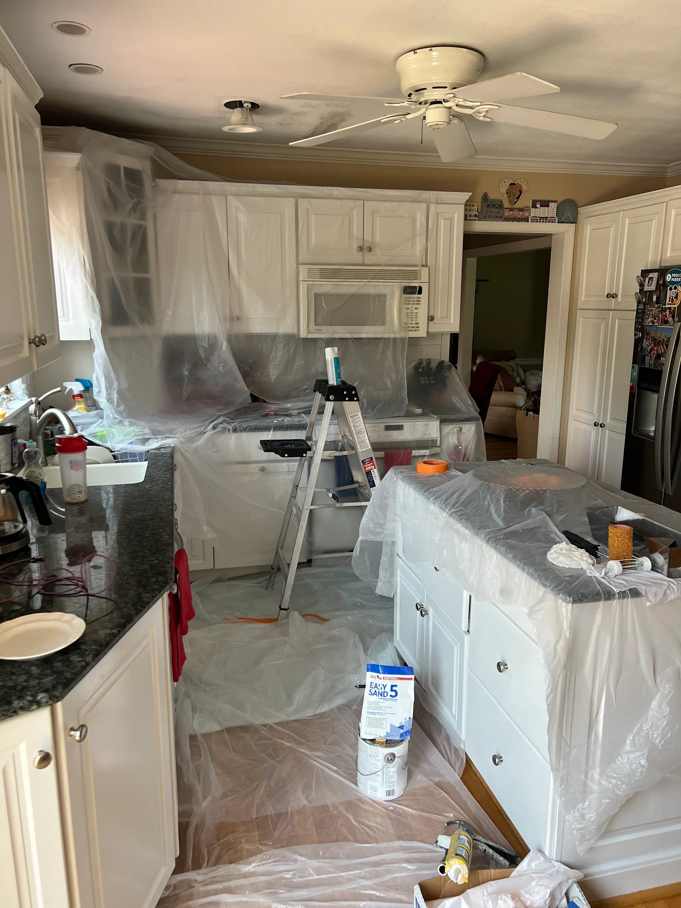 Kitchen prepped for ceiling drywall repair with plastic sheeting protecting cabinets and counters, joint compound bucket on the floor