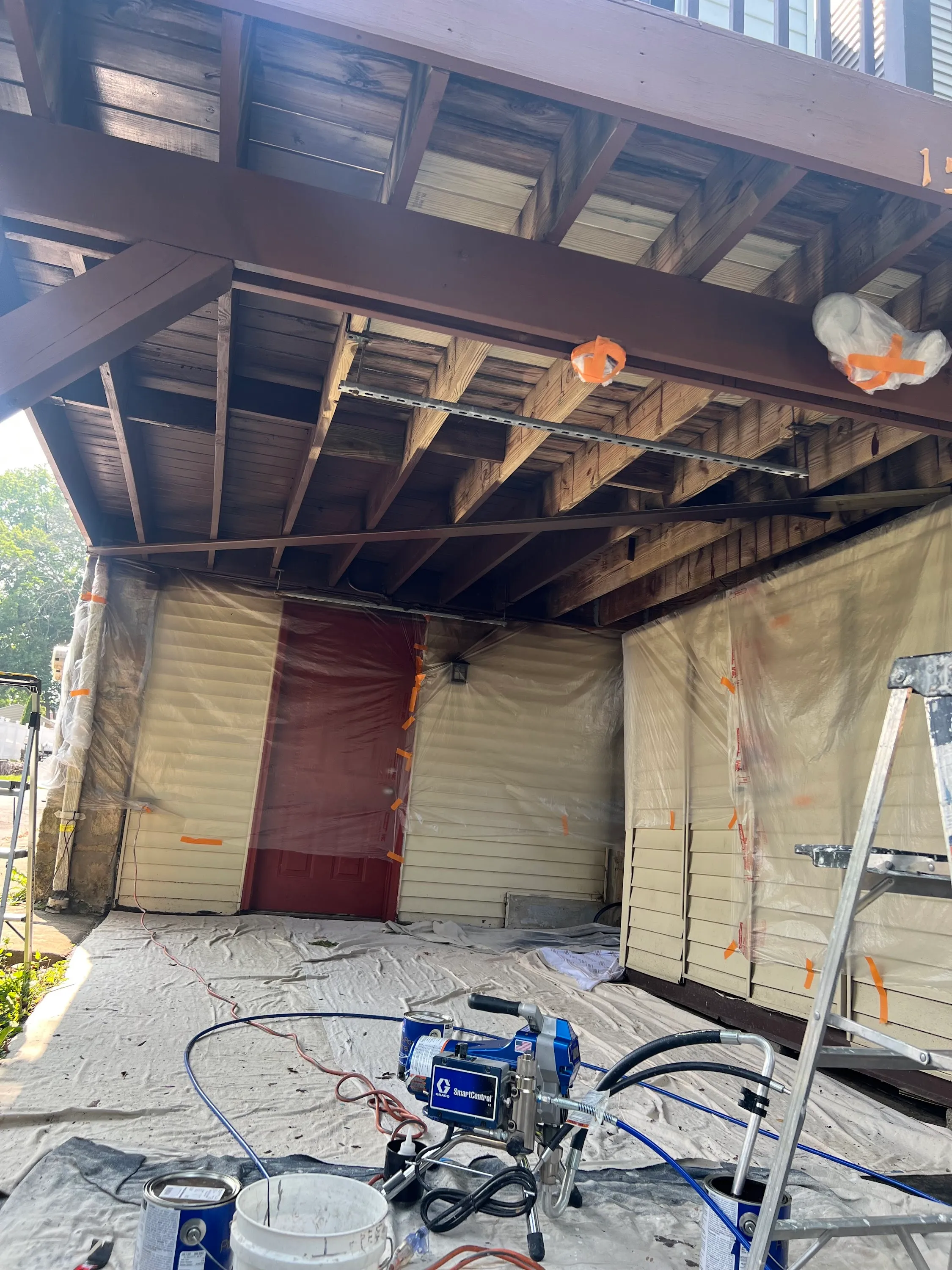 Pro-grade Graco airless paint sprayer set up on drop cloth under a wood porch ceiling with masking tape and plastic sheeting protecting the surrounding walls — the same containment discipline JL applies to popcorn-ceiling removal
